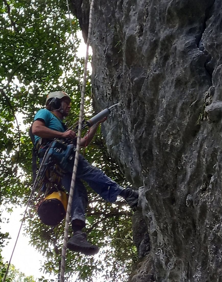 A volunteer in a helmet and ear muffs drilling into the rock while hanging on a rope